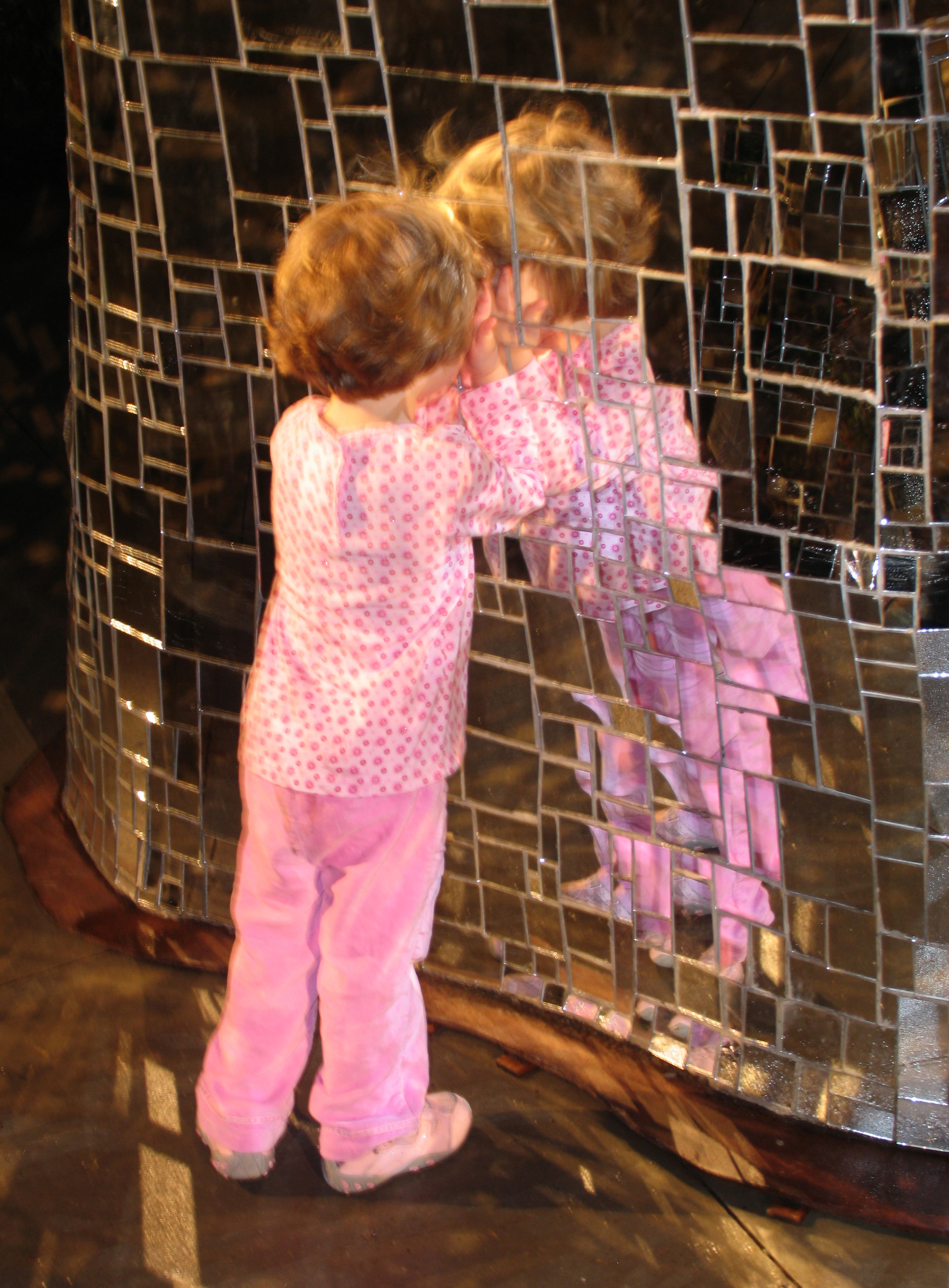 A young girl dressed in pink leans her forehead against a wall of glass tiles
